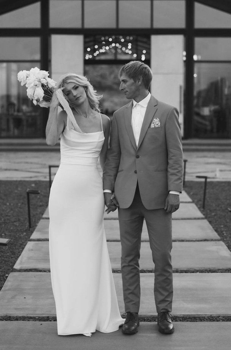 Bride in a crepe pleated bodice gown holds a bouquet and is standing beside her groom, on a path leading up to the venue.