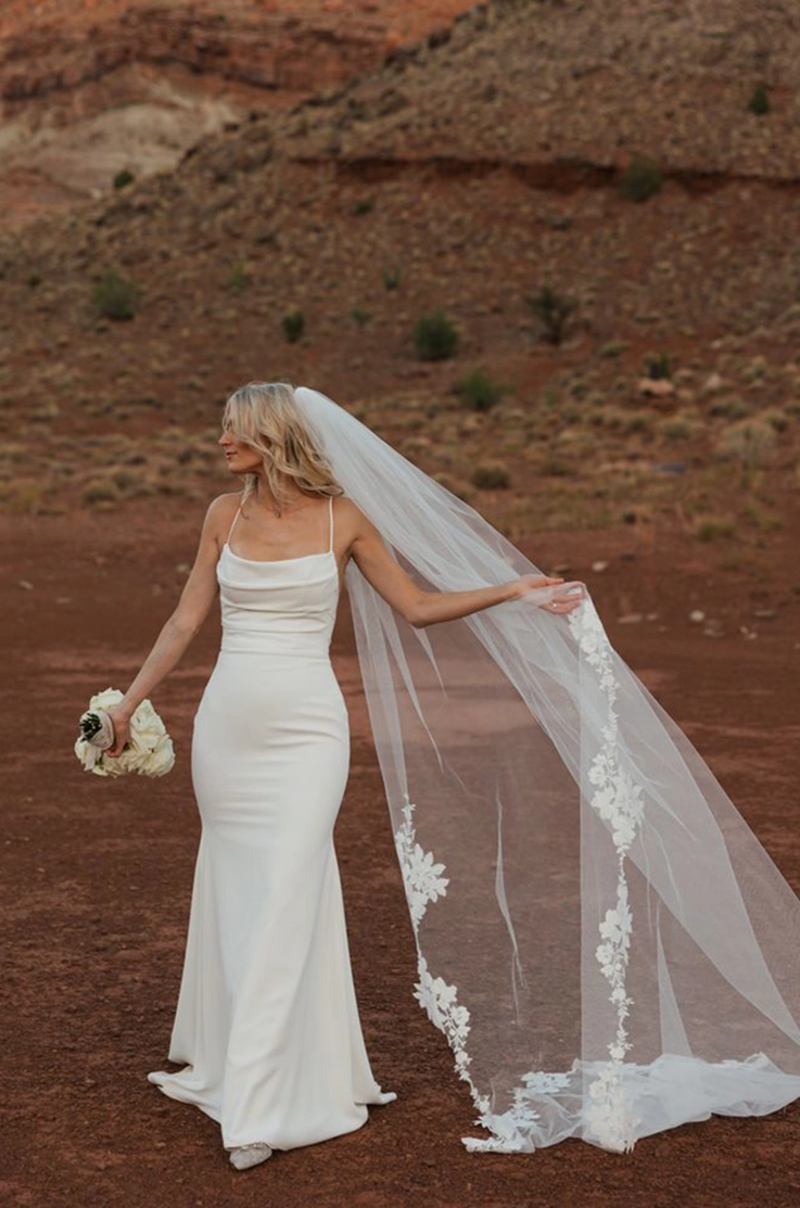 Bride in a fit and flare crepe gown with a pleated bodice and scoop neck, holds her veil and a bouquet in a canyon landscape. 