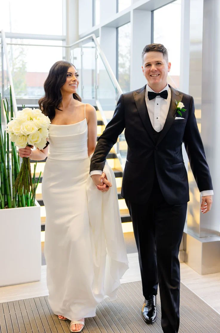 Bride in a crepe fit and flare gown with a pleated bodice, holds her train and a bouquet alongside groom at a staircase.