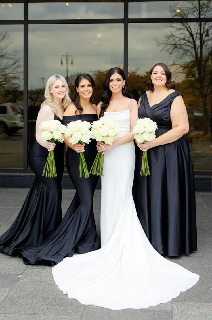 Bride in a sleek fit and flare crepe wedding dress holds a bouquet, next to her bridesmaids, outside the venue.