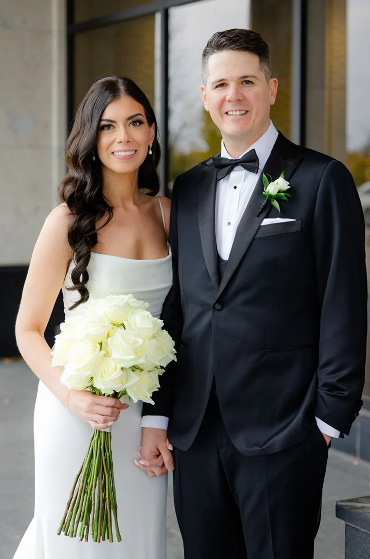 Close-up of bride in a fit and flare crepe gown with a scoop neck, is holding a white rose bouquet next to her groom.
