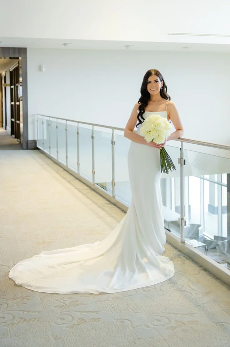 Bride smiles leaning on a railing in a crepe sleeveless gown with a scoop neckline, and is holding a white bouquet indoors.