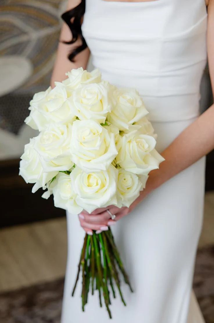 Close-up of bride in a fit and flare crepe gown with pleating, is holding a white rose bouquet indoors. 