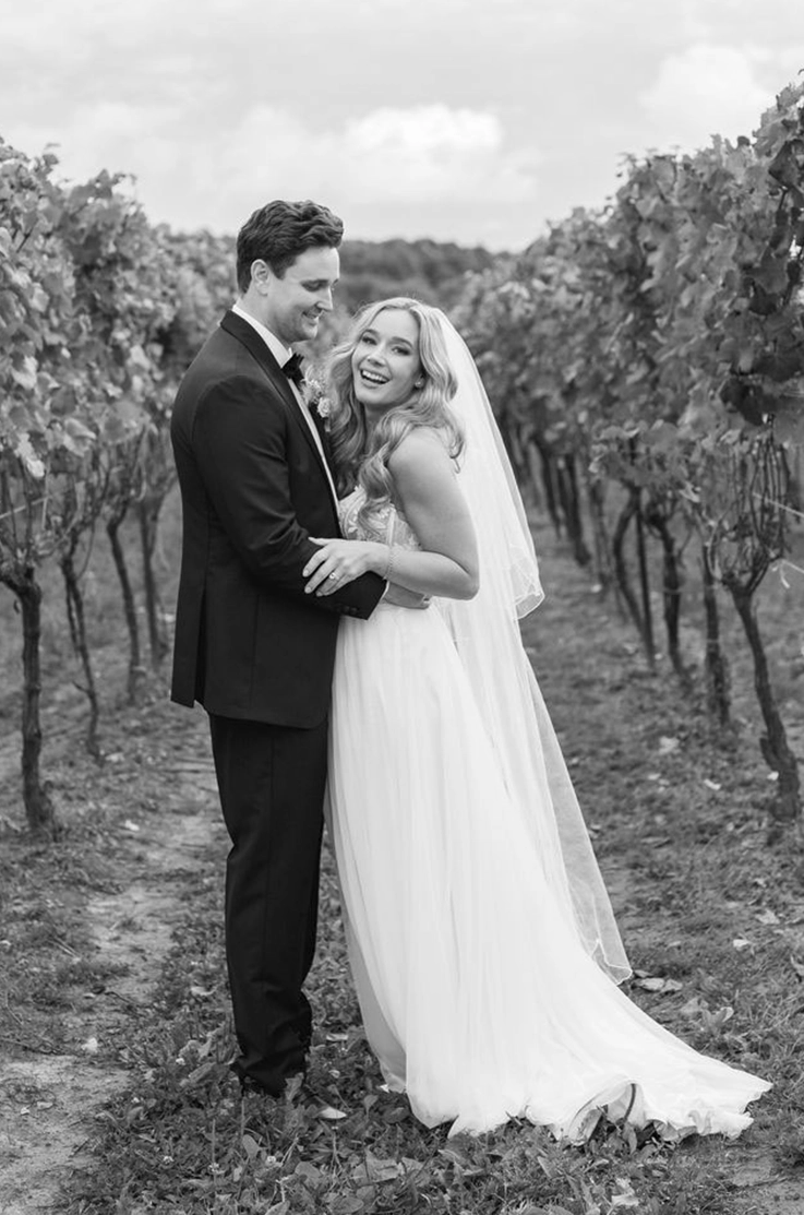  Bride in a lace bodice gown with a veil and tulle skirt, poses with groom in a vineyard row in black and white.