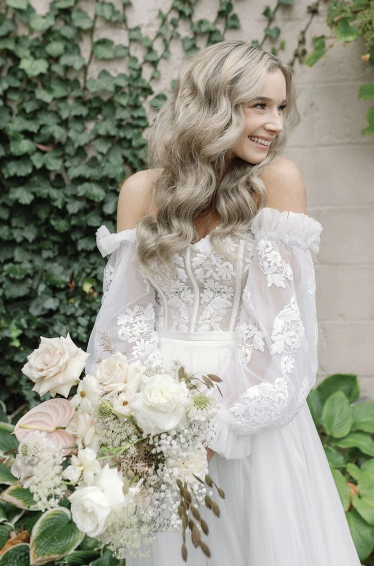 Smiling bride in a sheer lace bodice gown with sleeves, a fabric belt and tulle skirt, is holding a bouquet outdoors.