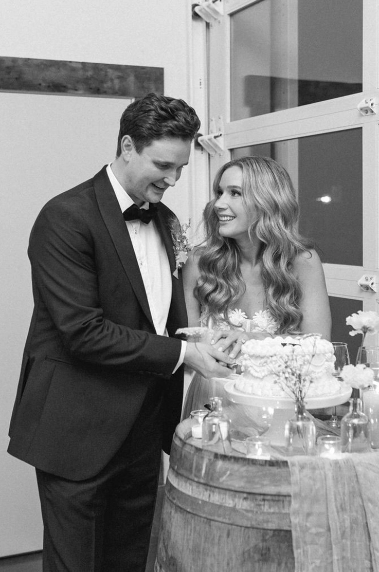 Bride in a strapless lace gown with a tulle skirt, smiles at groom by a garage door, as they cut cake in black and white.