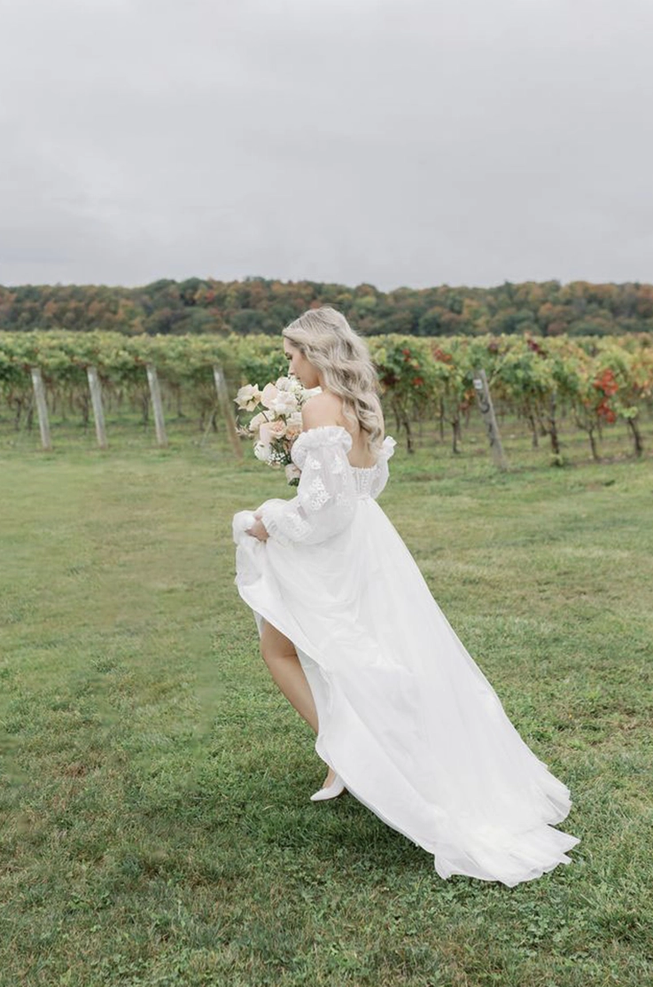 Bride in an off-shoulder lace and tulle wedding dress, is holding a blush bouquet walking through a vineyard.