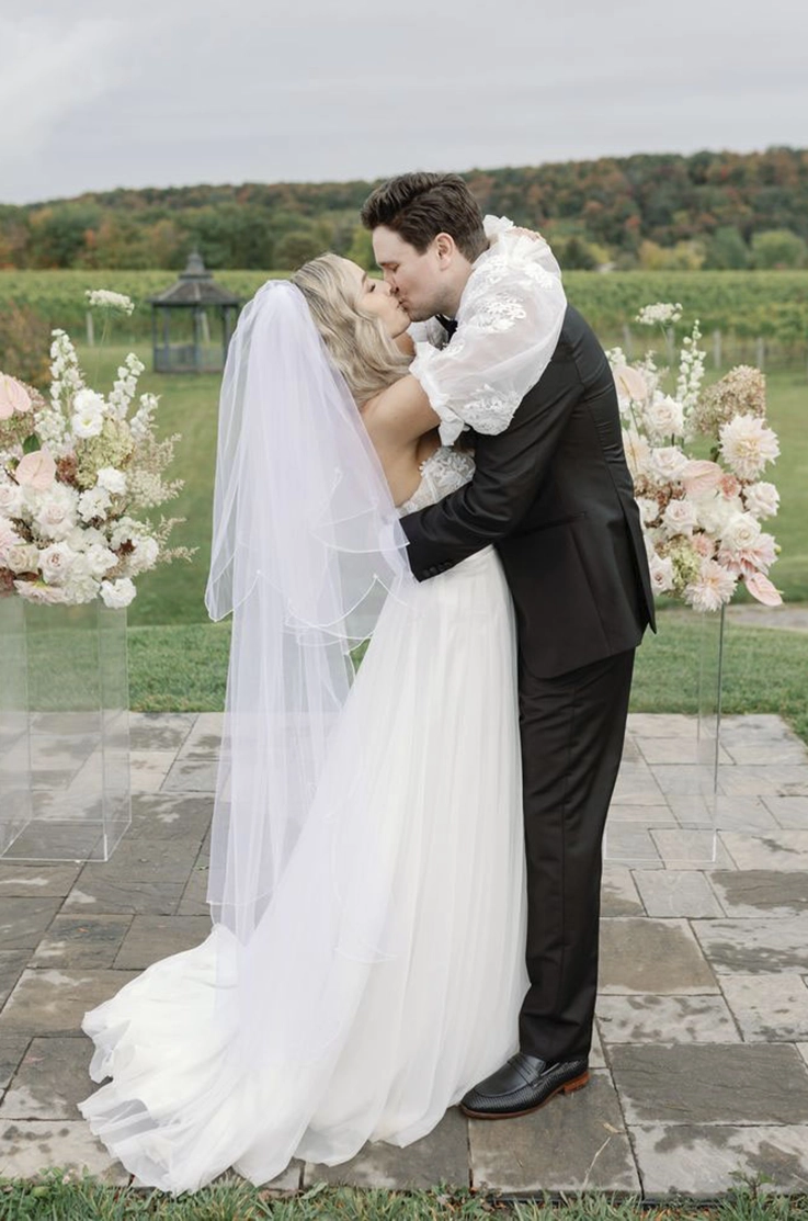 Bride in a strapless lace and tulle gown with off shoulder sleeves, kisses her groom between two flower posts at a vineyard.