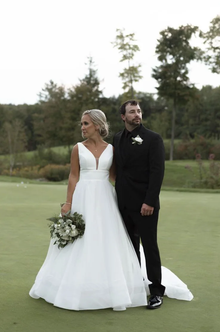 Bride in a sleeveless crepe and organza wedding dress poses with her groom, holding a bouquet on a golf course.