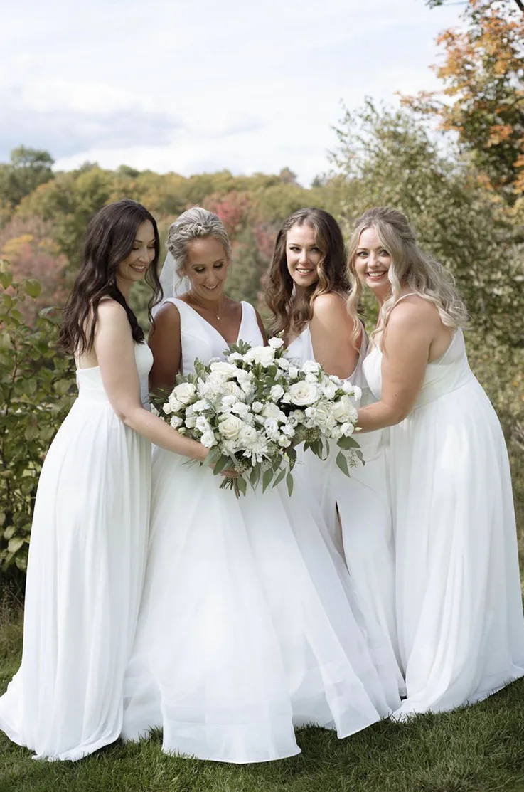 Bride in a V-neck crepe gown with an organza skirt, poses with bridesmaids holding white bouquets together by autumn trees