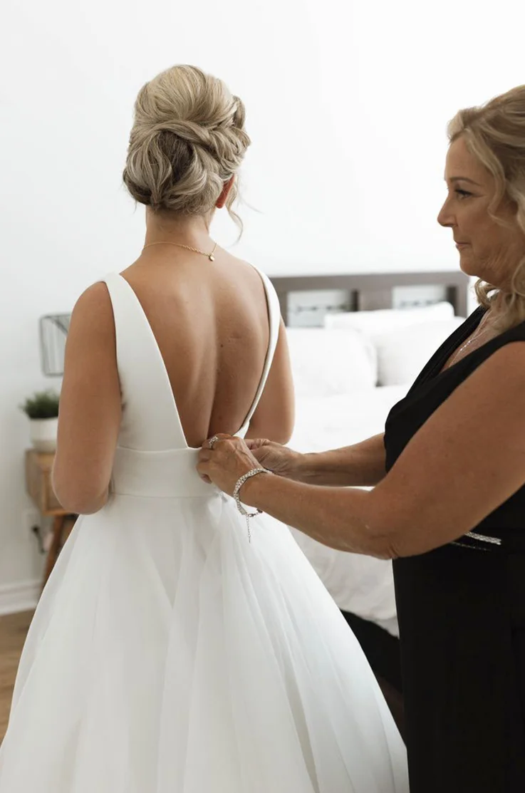 Bride is fastened into a crepe bodice gown with an empire waist and organza, by her mother in a black dress in a bridal suite. 