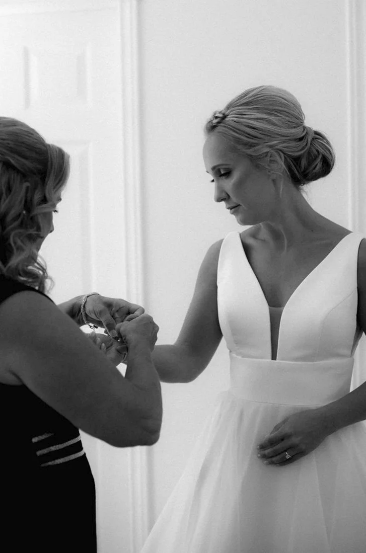 Close-up of bride in a plunging neck crepe gown with an organza skirt, is getting ready indoors in black and white. 