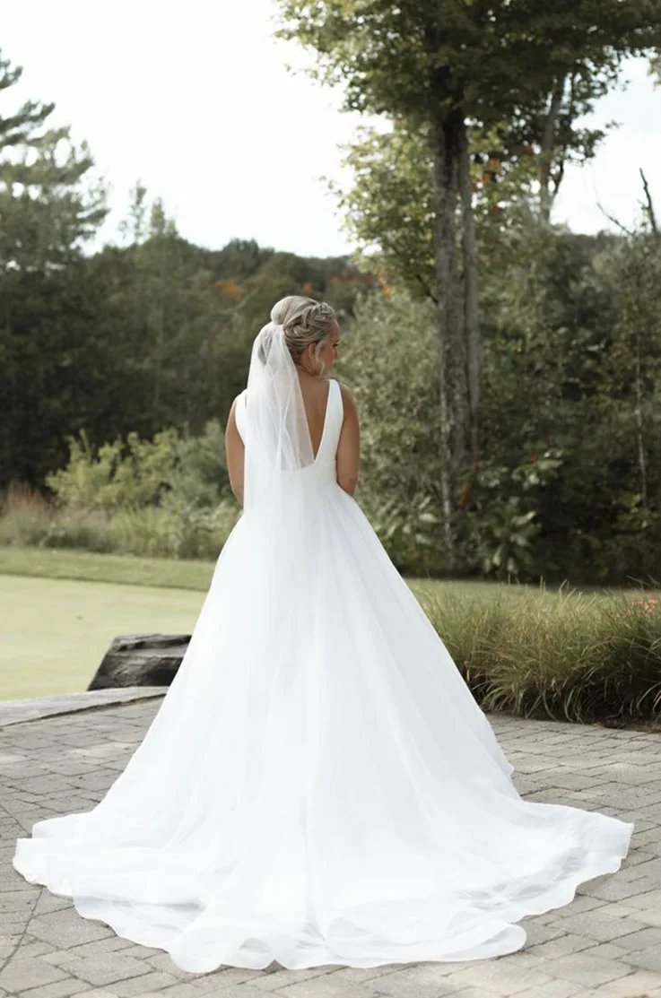Back of bride, posing in a crepe and organza gown with a veil and a low back, is standing on a stone patio near trees. 