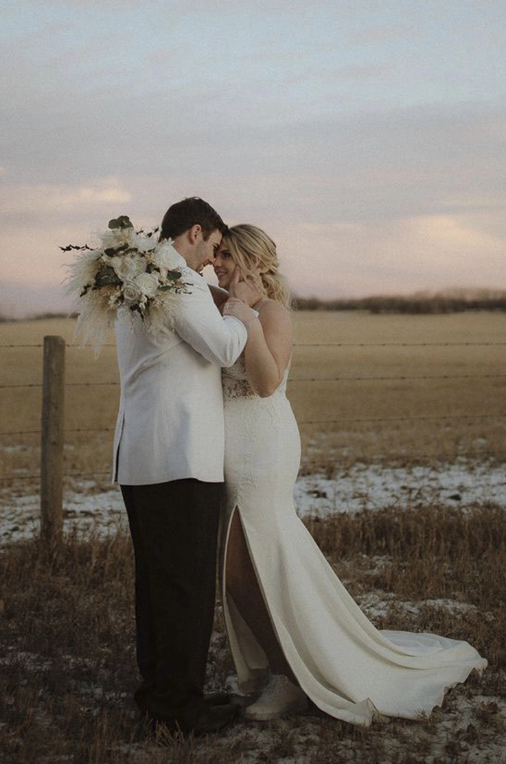 Bride in a fit and flare crepe gown with lace and a skirt slit, is holding a bouquet and faces her groom in an open field