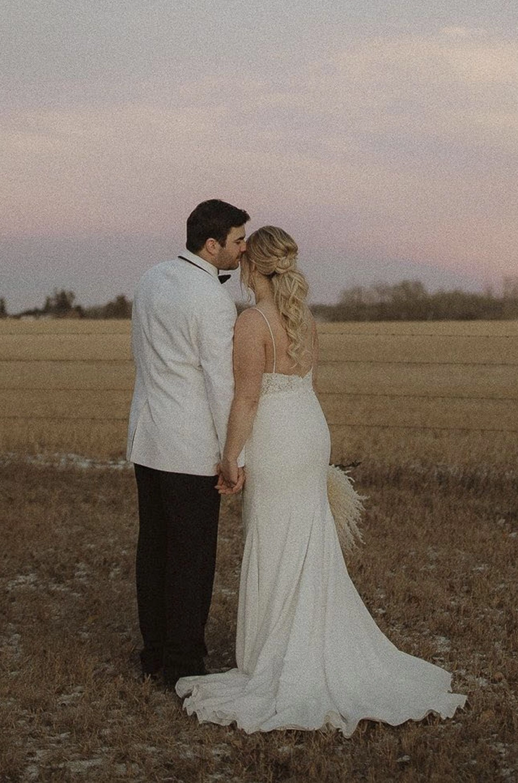 Bride in a fitted lace and crepe gown with a low back and thin straps, poses with groom in an open golden field.