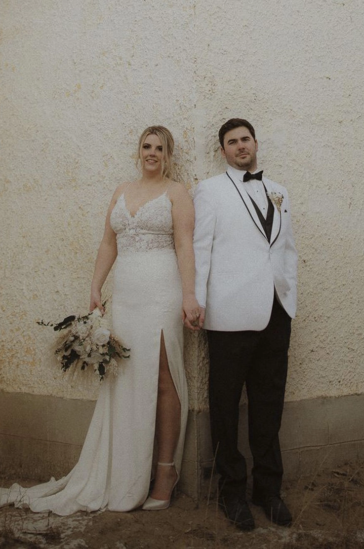 Bride in a fit and flare lace bodice wedding dress with a crepe skirt and slit, poses next to groom at a white wall