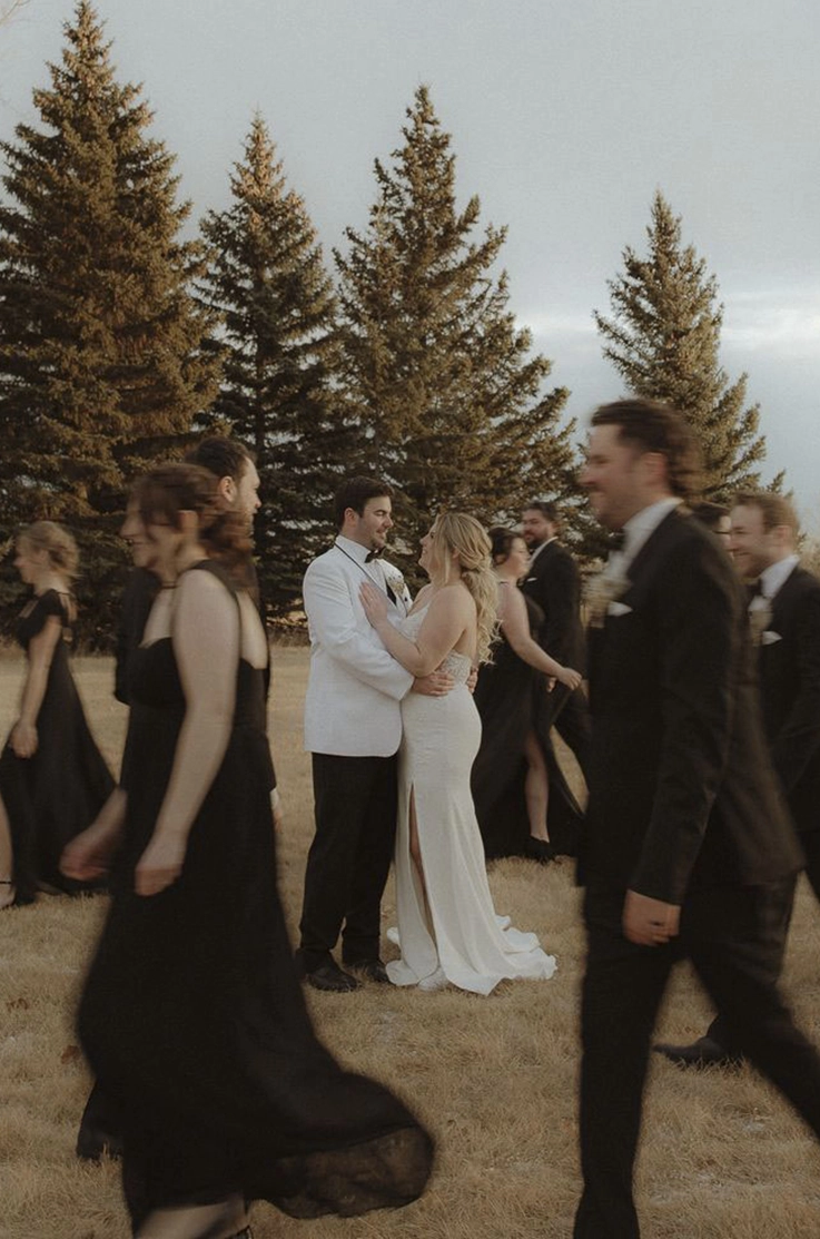 Groom holds bride in an embrace in a crepe gown with a skirt slit and V-neckline, surrounded by bridal party in black. 
