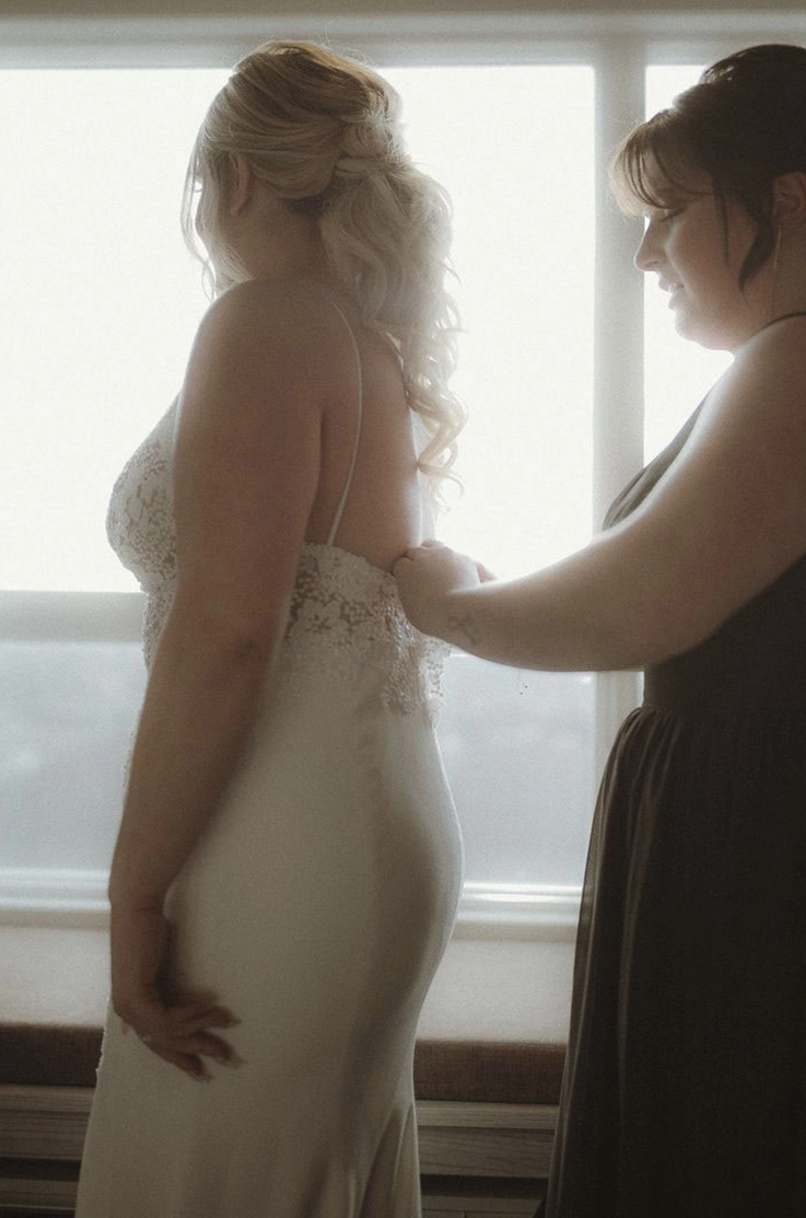 Bride gets ready in a sleeveless lace gown with a fit and flare crepe skirt, by a bright window, as a bridesmaid helps