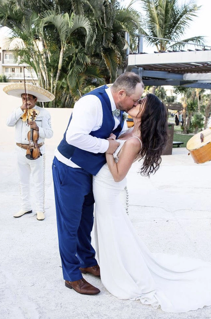 Groom kisses his bride in a lace bodice gown with a fit and flare crepe skirt, in front of a mariachi band and palm trees.
