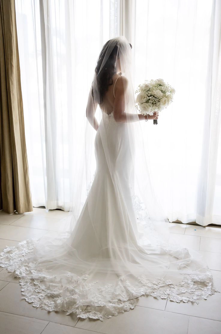 ride in a crepe fit and flare gown with a lace-trim veil, is holding a bouquet and faces a window with white curtains.