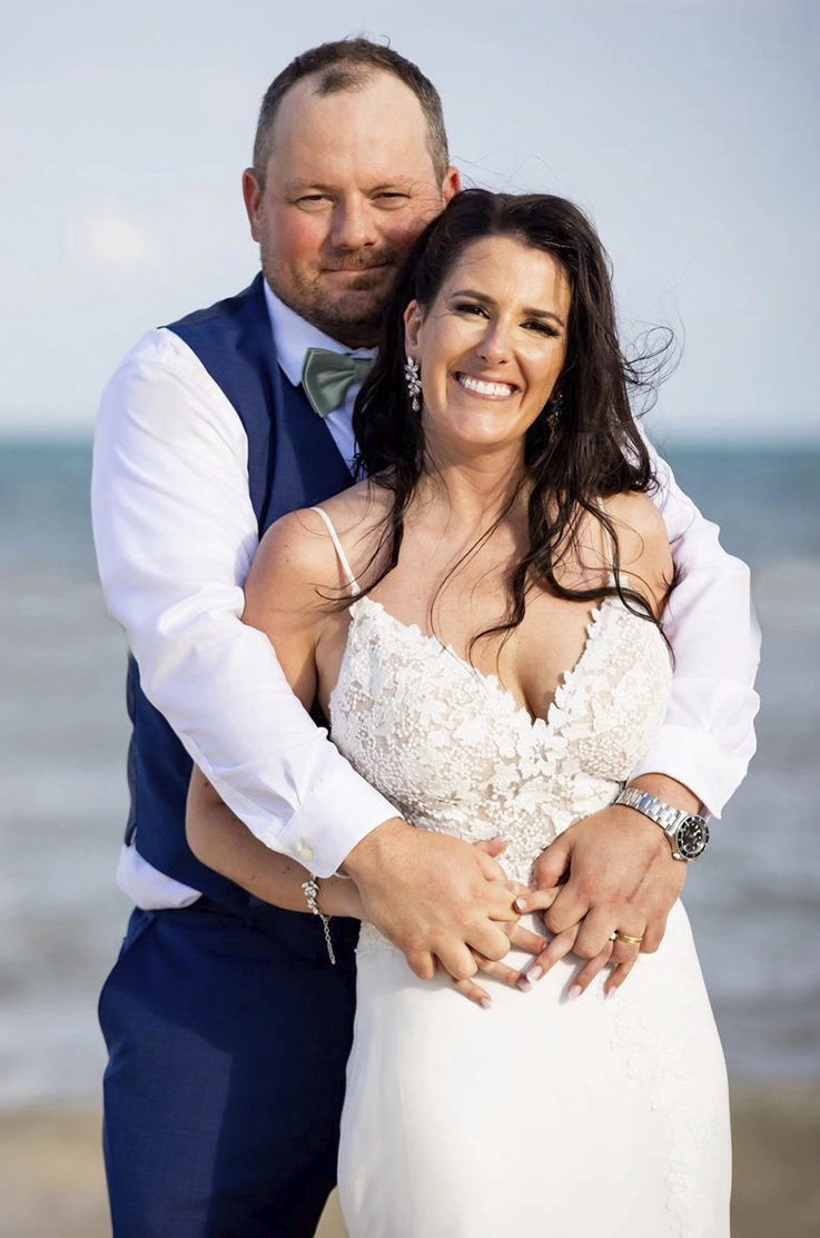 Groom holds his bride in an embrace, on a beach in a V-neck lace gown with a crepe skirt and thin straps. 