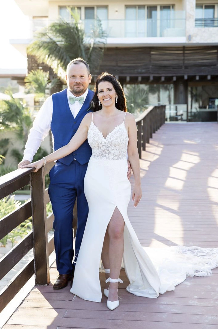 Bride in a V-neck lace and crepe wedding dress with a skirt slit, poses beside her groom at a resort on a bridge.