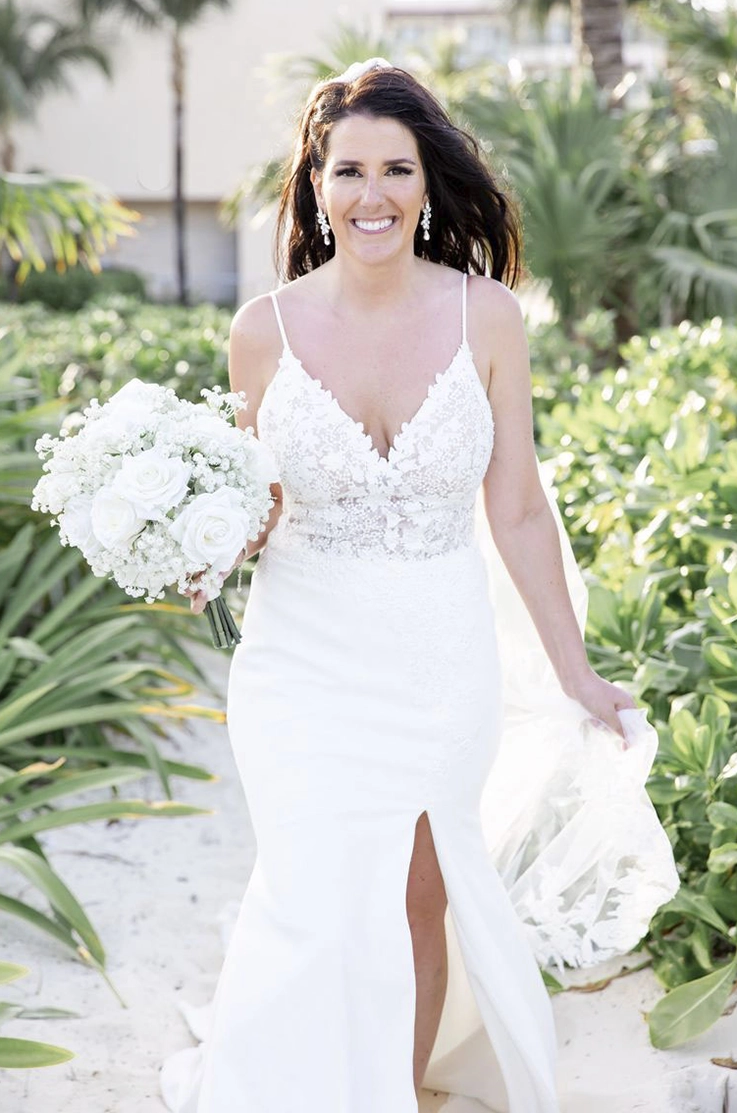 ride in a sleeveless lace bodice gown with a V-neck and fitted crepe skirt with a slit, is holding a white bouquet outdoors. 