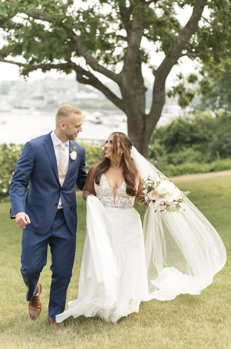 Bride in a sleeveless tulle gown with a lace applique bodice, holds her train and walks alongside the groom.