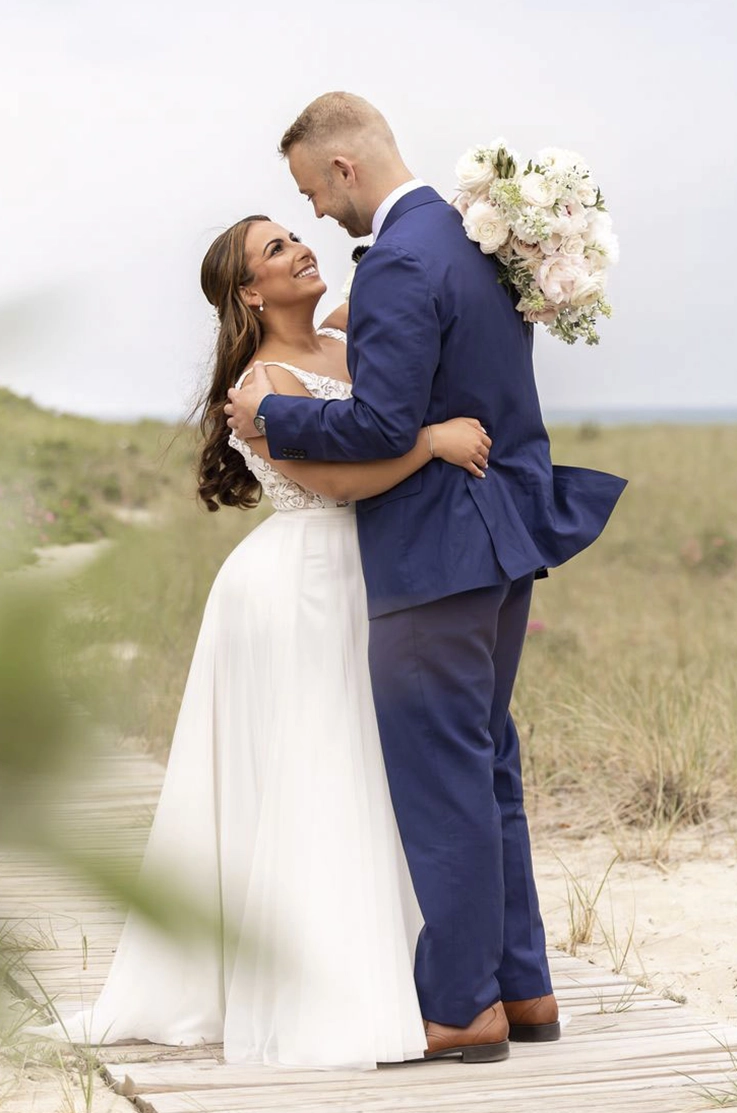 Groom embraces his bride in a sleeveless lace and tulle wedding dress, holding a bouquet on a boardwalk at a beach.