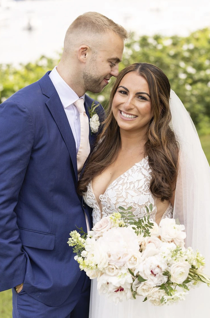 Close-up of bride in a V-neck embroidered tulle and lace gown with a full tulle skirt and a veil, smiling beside her groom.  
