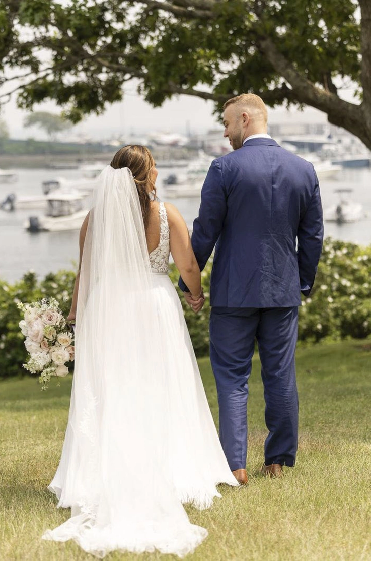 Bride in a lace bodice and flowing tulle gown with a veil, poses next to her groom, and looking out at a sea with boats. 
