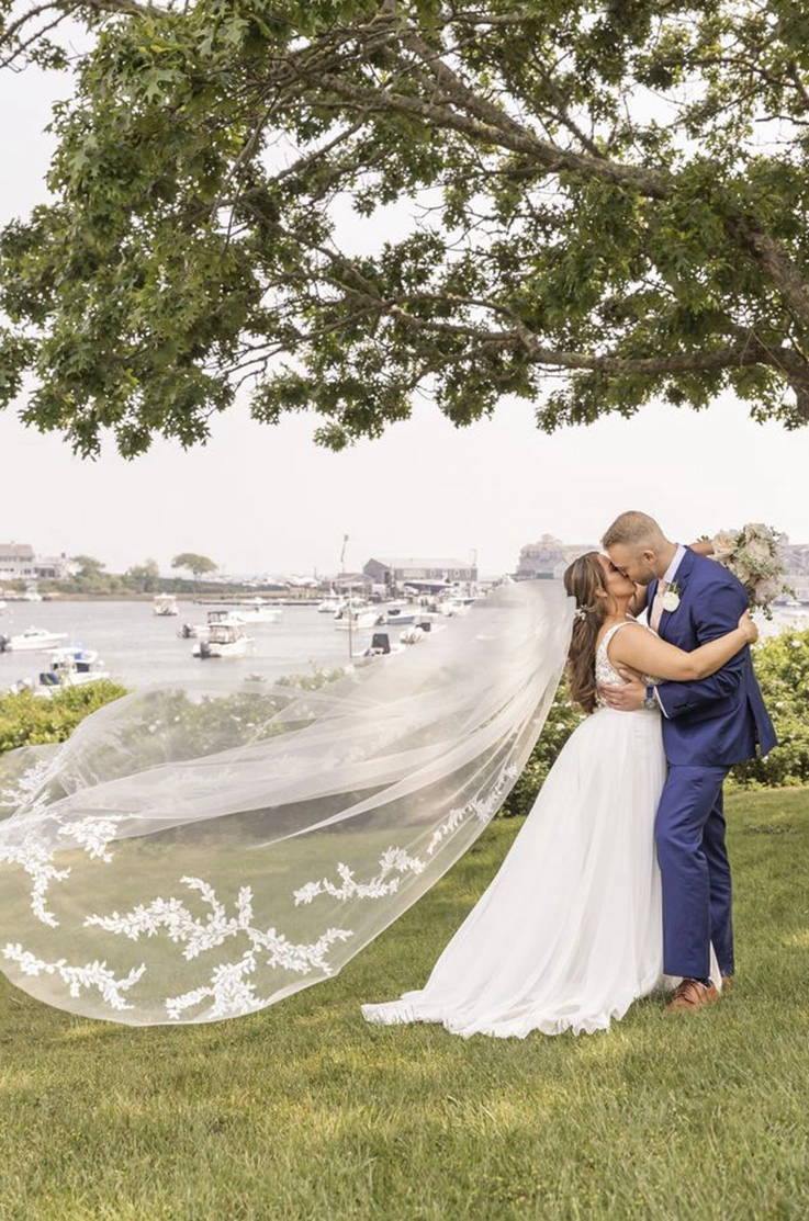 Groom embraces bride in a tulle A-line gown with a lace-trim veil blowing in the wind, under a large tree by a waterfront.