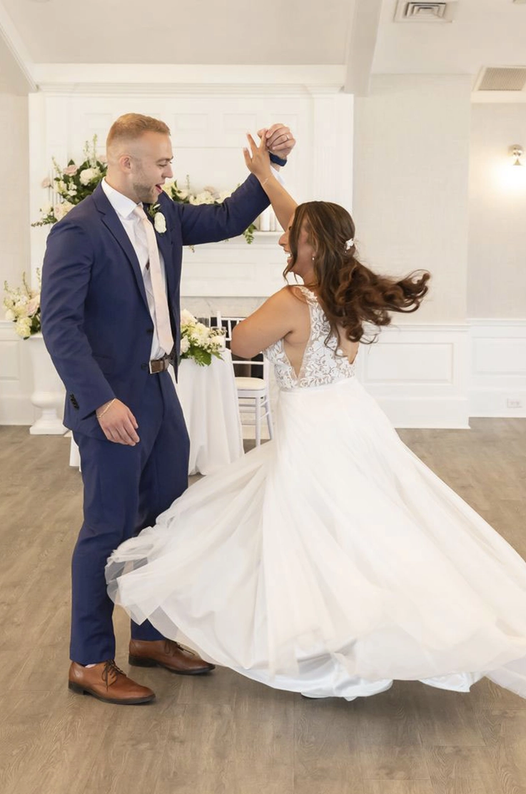 Groom in a blue suit twirls his bride in a lace bodice and tulle gown with side cutouts, in an elegant ballroom