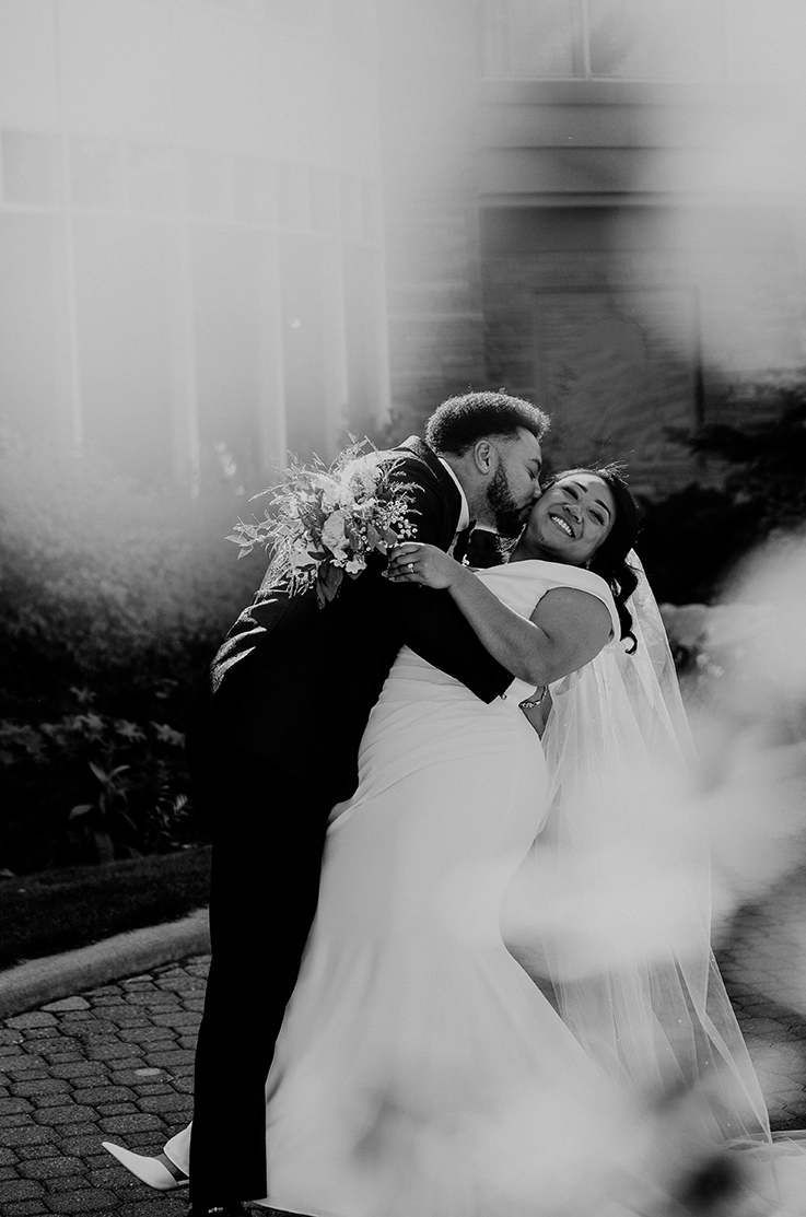 Groom dips his bride and kisses her cheek in a crepe fit and flare gown with a draped bodice and veil, in black and white.