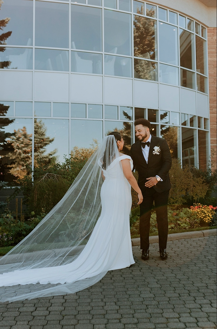 Bride in an off-shoulder V-shaped back wedding dress with a flowing veil, faces her groom outside the venue.