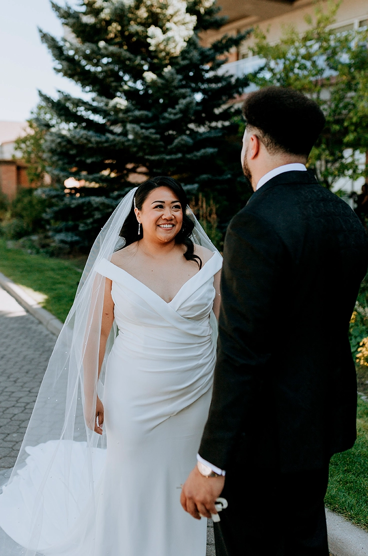 Groom faces bride in a draped crepe bodice gown with an off-shoulder neckline and a veil, on a sunlight path lined with trees.