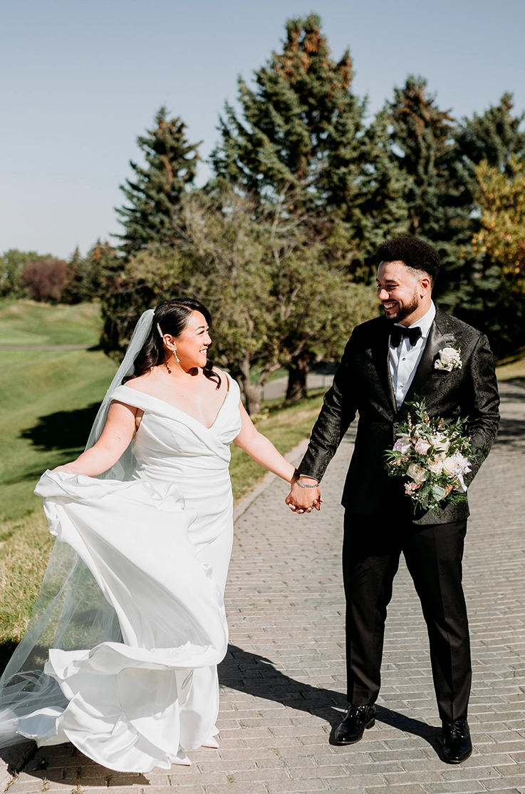 Bride in an off-shoulder V-neck crepe gown with draping and a veil, holds her train and poses with her groom on a stone path.