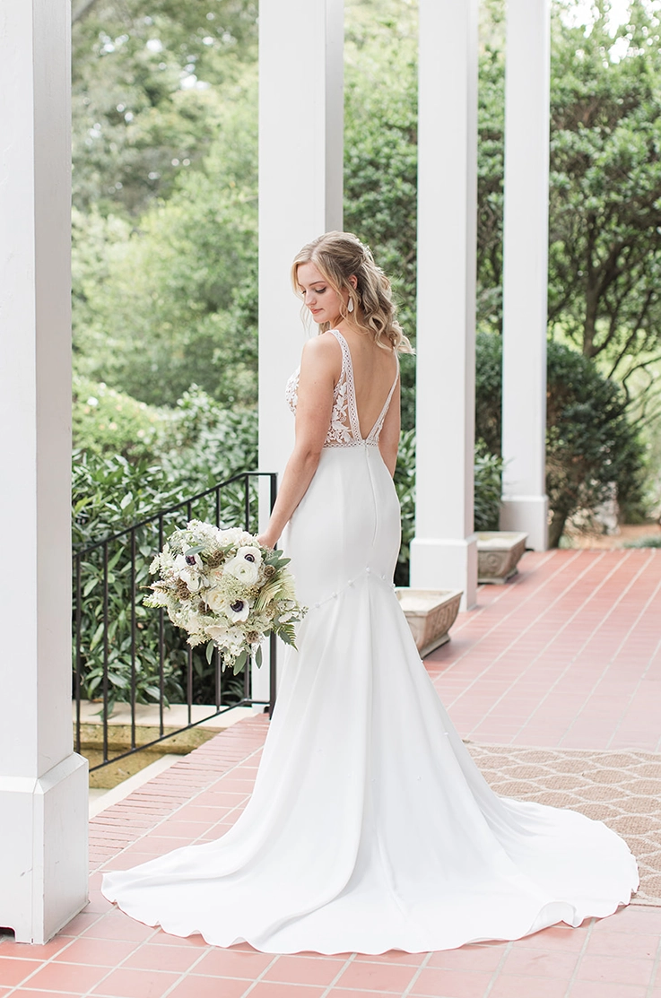 Bride poses in a deep-V back lace gown with a crepe fit and flare skirt, is holding a large bouquet on a veranda. 
