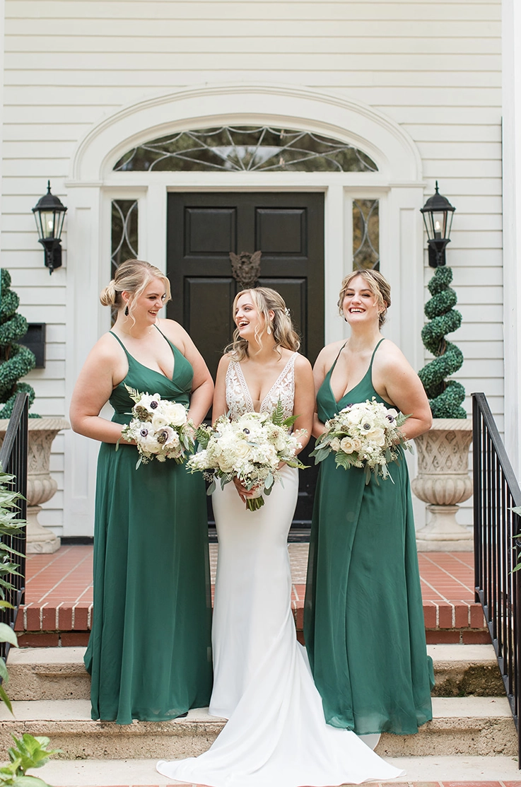 Bride in a lace bodice wedding dress with a crepe skirt, poses between her bridesmaids in green dresses, at the venue doors.