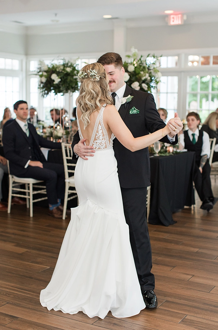 Bride in a sleeveless lace gown with a crepe fit and flare skirt, dances with her groom in a ballroom in front of guests. 