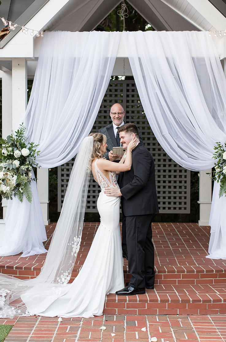 Bride in a sleeveless lace gown with side cutouts, a crepe fit and flare skirt and veil embraces groom at the altar. 