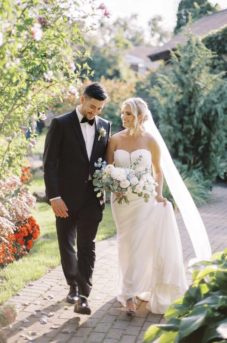 Bride in a lace sweetheart neck gown with a crepe fit and flare skirt and a veil, walks alongside her groom on a garden path.