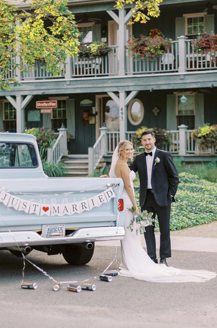 Bride in a strapless fit and flare wedding dress leans against a vintage truck, beside her groom and in front of the venue.