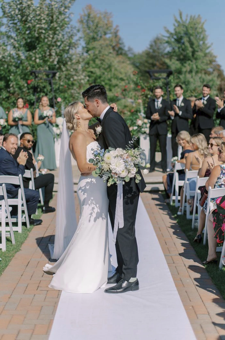 Groom kisses his bride in a fit and flare crepe gown with lace on the bodice and a veil, holding a bouquet in the aisle.