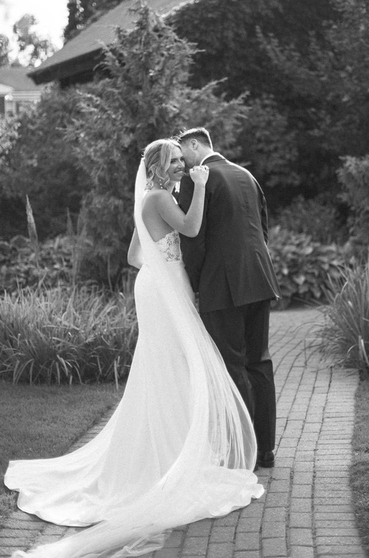 Bride in a lace bodice and crepe flowing gown with a long veil, poses beside her groom on a garden path in black and white. 