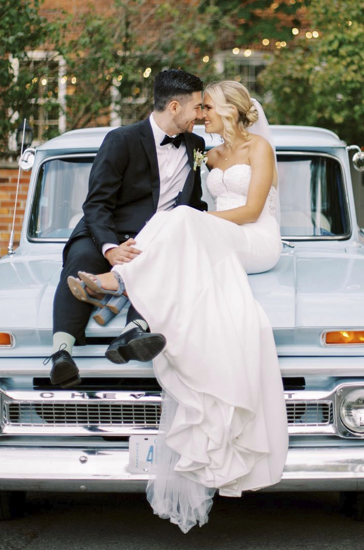 Bride in a strapless crepe gown with a sweetheart neckline and a veil, poses with her groom on a blue vintage car. 