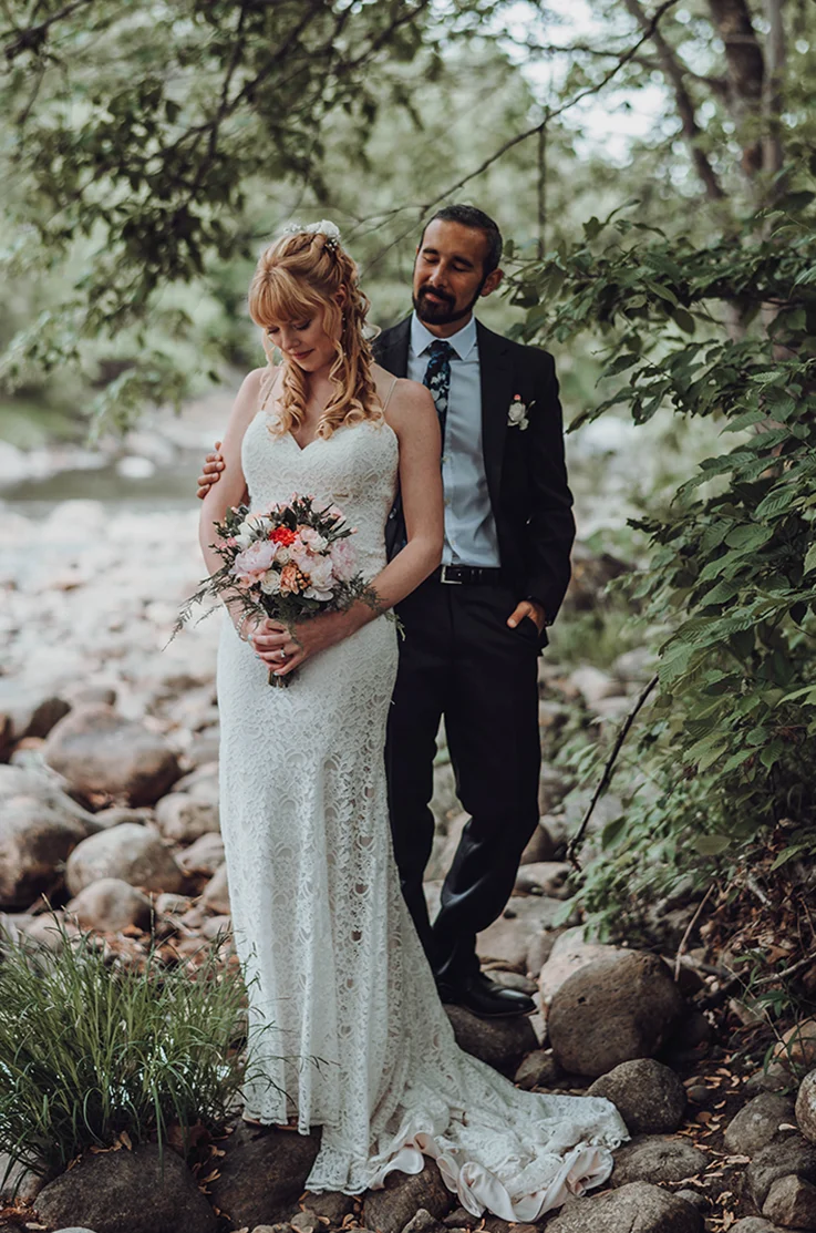 Bride in a fit and flare lace wedding dress, is holding a bouquet next to her groom by a riverside stream in a forest.