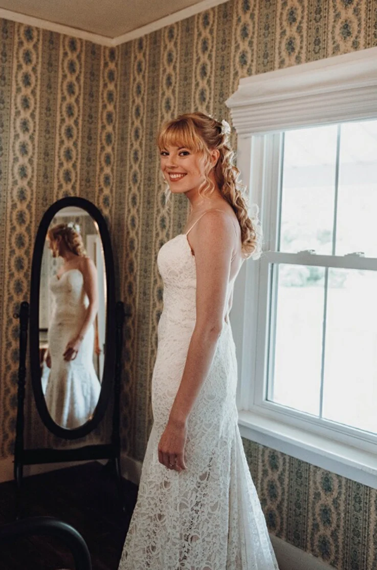 Bride smiles in a sweatheart neck lace gown, and poses in a mirror in a brightly lit room, next to an open window.