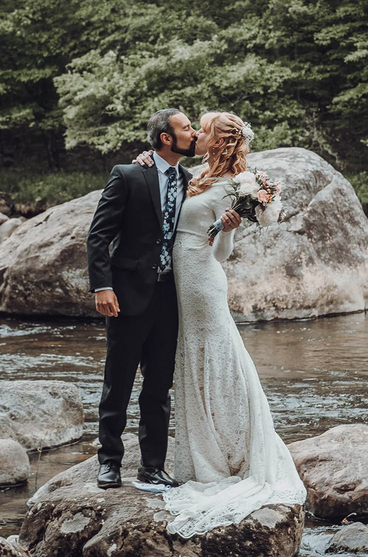 Groom kisses bride in a lace fit and flare gown with sleeves, holding a bouquet as they stand on rocks in a stream.