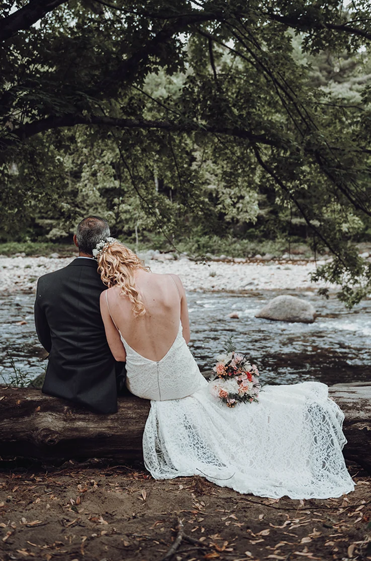 Bride in a deep-V back lace gown, sits next to groom on a wood log looking out at a river stream, and are surrounded by trees.